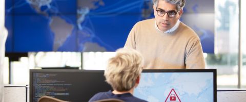 An IT security professional stands facing a colleague seated at a desk with two computer monitors, one displaying code and the other showing a red lock icon inside a triangle. In the background, there are large screens displaying a world map with network connections highlighted.