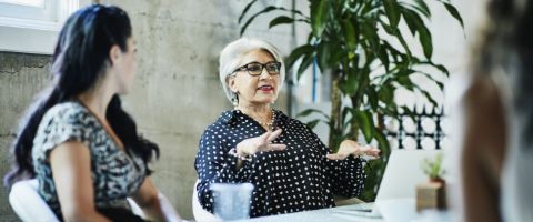 Two individuals sit at an office table, as one woman speaks to the those around her and the other listens.