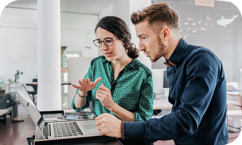 Two businesspeople sit in front of a laptop in an office setting and engage in discussion. 
