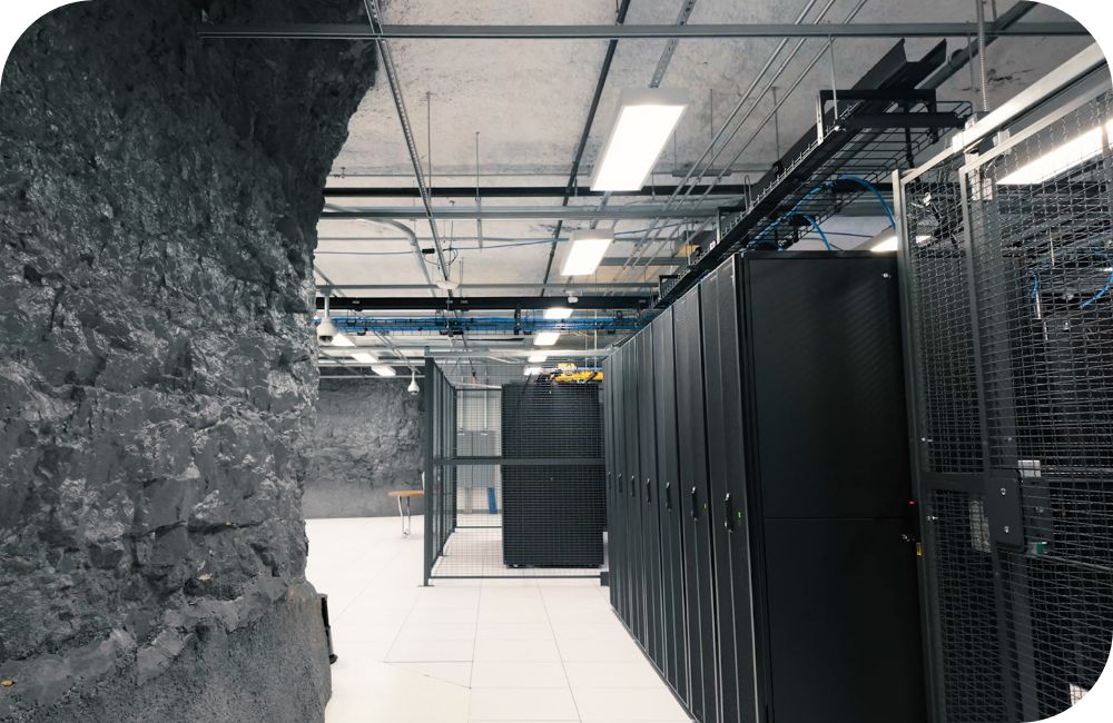 Interior of a modern data center with rows of black server cabinets enclosed in metal mesh, visible cable trays and wiring overhead, bright fluorescent lighting and rough stone walls on one side.