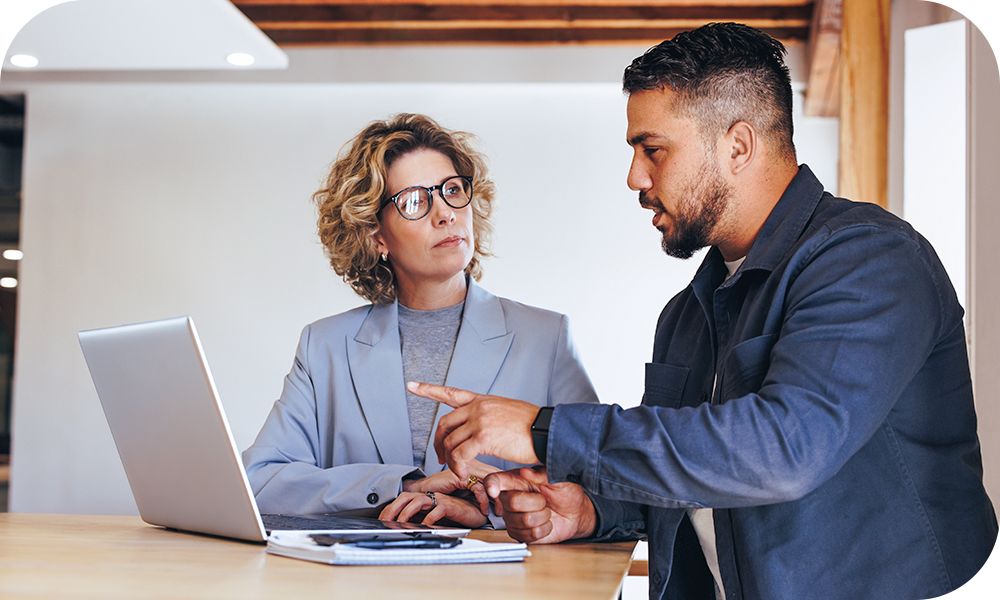 Two professionals chat while one gestures at a laptop in front of them and the other listens.