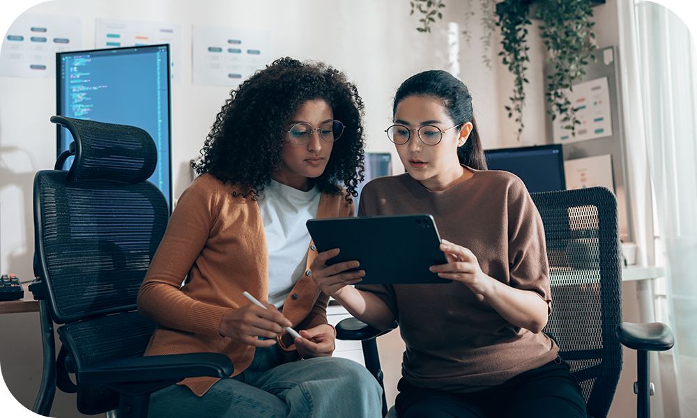 Two colleagues sitting in desk chairs look at a tablet together.
