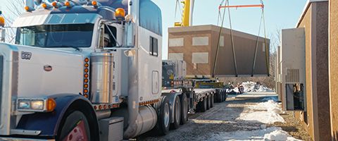 Flatbed truck transporting a large prefabricated concrete structure being lifted by a crane at an outdoor construction site with snow on the ground.