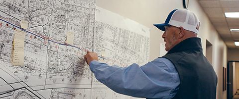 Person standing indoors and pointing to a large wall mounted map or schematic, with detailed street lines and markings visible across the surface.