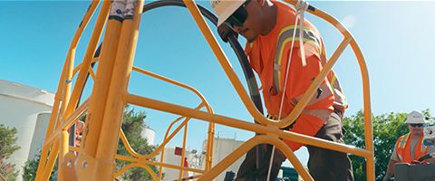 Person wearing high visibility safety gear operating equipment on a yellow metal structure at an outdoor industrial site under a clear blue sky.