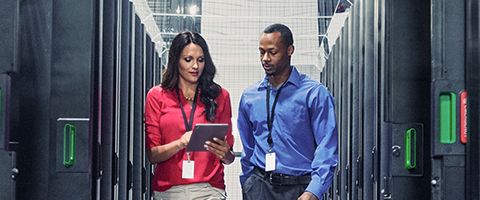 Two IT professionals stand in a data center aisle, looking at a tablet between rows of server racks.