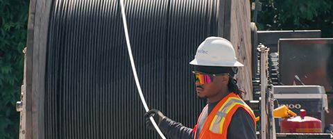 A worker in safety gear stands beside a large industrial cable spool, handling a cable line at an outdoor worksite.