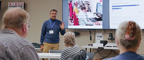 A presenter stands at the front of a room addressing seated attendees, with a large display behind them showing people working at computers.