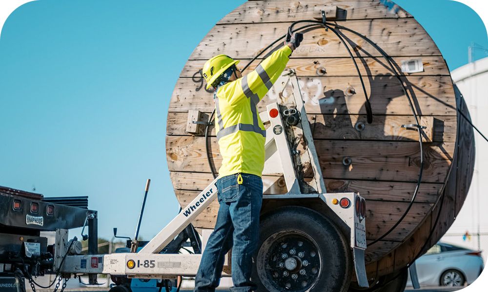Person in high visibility safety gear unloading or securing cable from a large wooden spool mounted on a trailer at an outdoor work site under a clear sky.