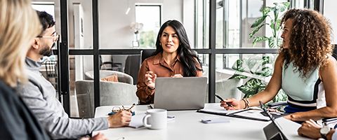 Person seated at a table in a modern glass walled office, working on a laptop while gesturing during a discussion, with other participants’ hands and documents visible in the foreground.