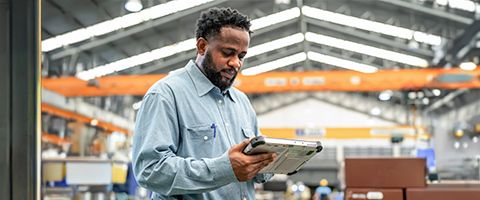 Person standing inside a large industrial facility, reviewing information on a tablet with structural beams and equipment visible in the background.