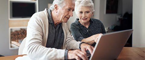 Two older adults seated at a table indoors, working together on a laptop, with one person typing and the other pointing toward the screen.