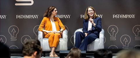 Two speakers seated on stage during a panel discussion, sitting in armchairs in front of a branded backdrop at a professional conference or live event.