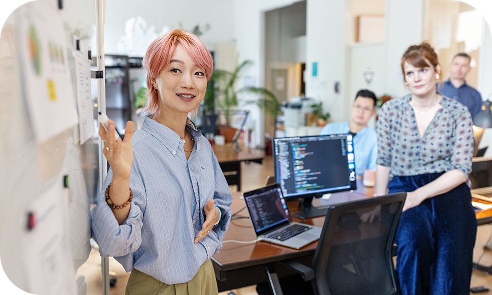 Woman with pink hair presenting in front of three other people sitting at a row of desks and laptops.