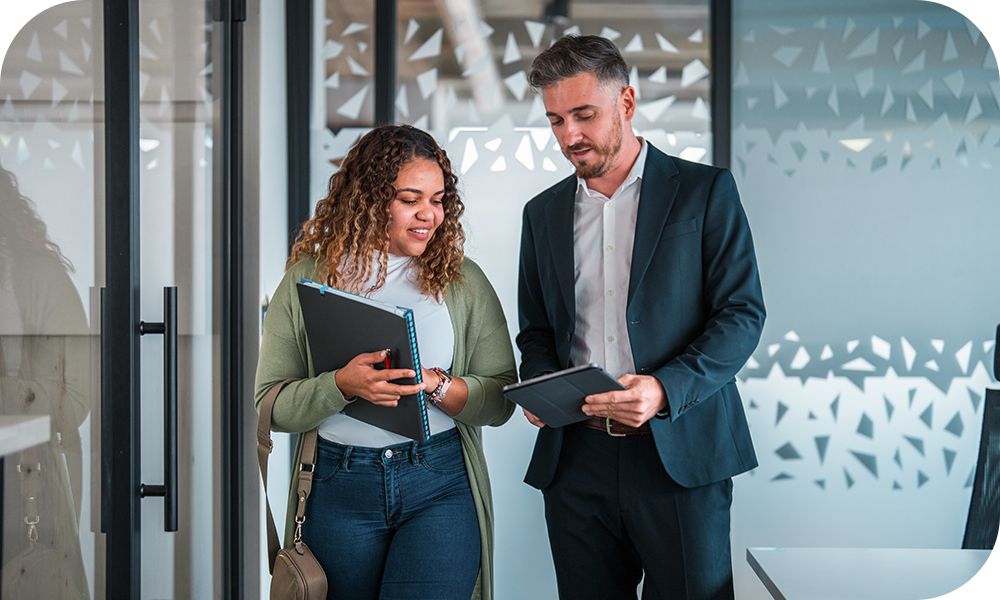 Two businesspeople walk side-by-side looking at the tablet in one of their hands. 