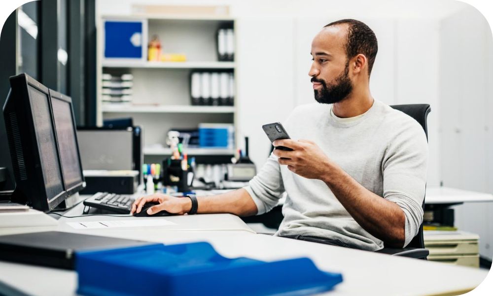 An individual works at a desk while looking at a monitor and holding a cellphone.