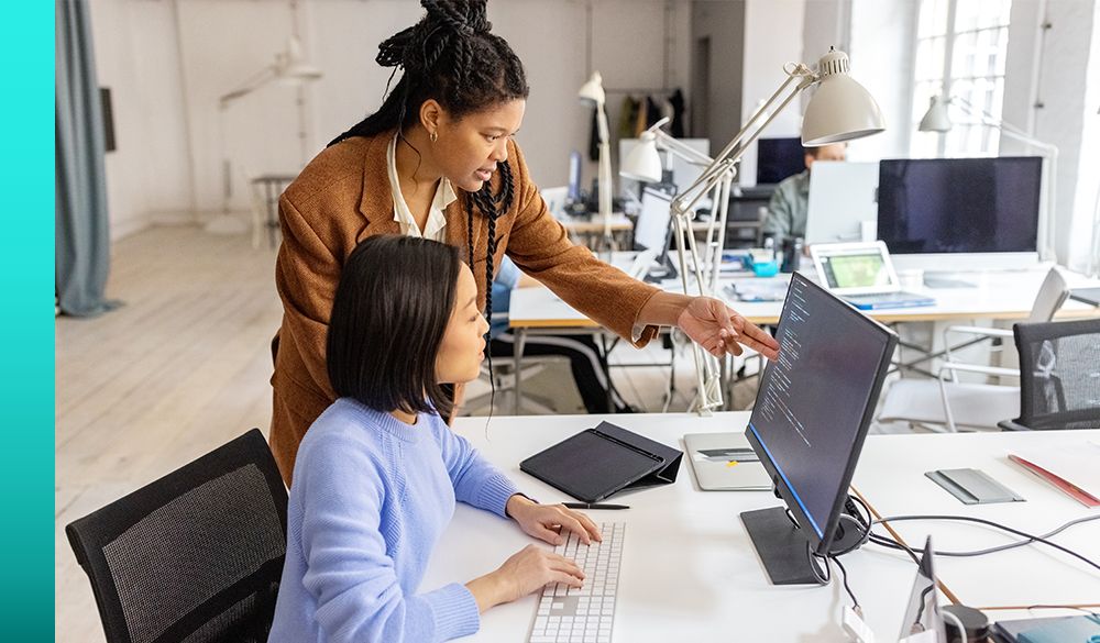 Two businesspeople have a professional discussion, with one sitting at their desk typing and the other pointing at a computer before them both showing code. 