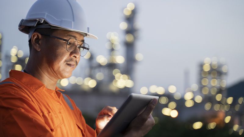 Energy worker in protective uniform and helmet uses a tablet device with energy plant infrastructure in the background