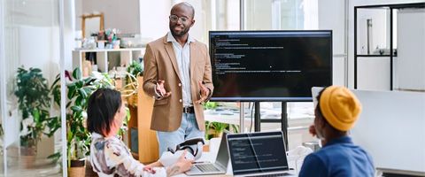 A person in business casual attire giving a presentation in front of a monitor to seated coworkers in an office.