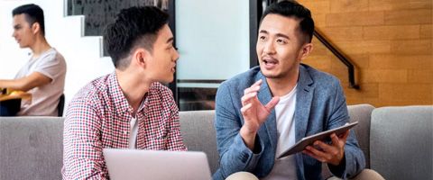 Two people in casual attire sitting on a couch holding a tablet and laptop while talking.