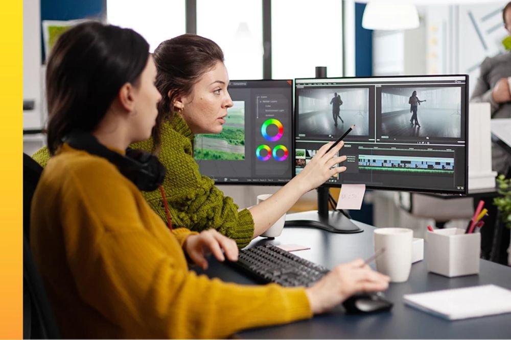 Two people work at a desk in a video editing studio, reviewing footage on dual monitors. One monitor shows a video editing timeline and preview windows, while a smaller screen displays color correction tools.