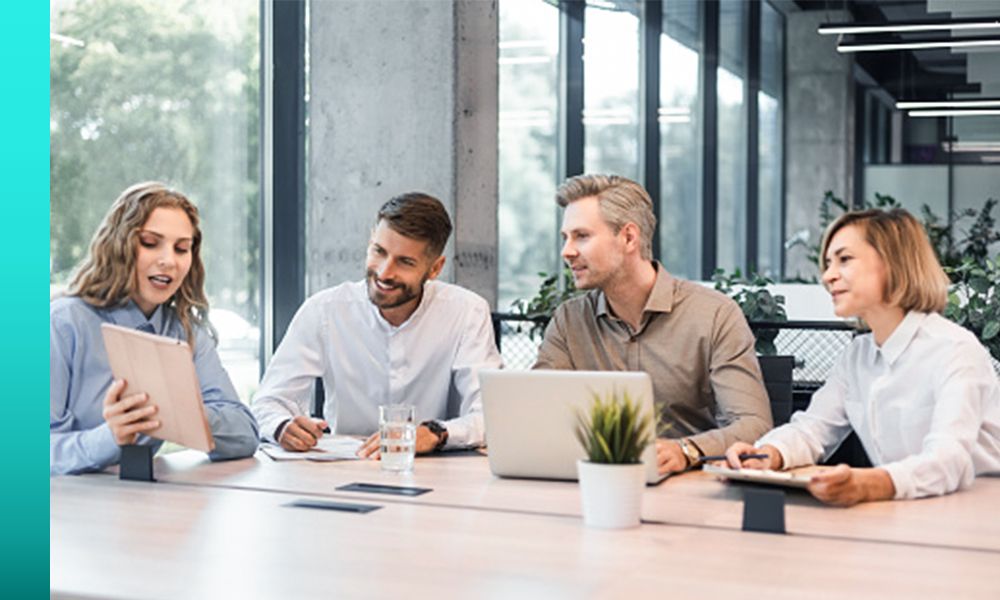 Four colleagues sit around a wooden conference table in a modern office with large windows and industrial concrete columns, reviewing content displayed on a tablet.