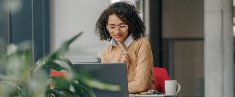 Businessperson in a tan sweater and glasses reads something on a laptop screen. 