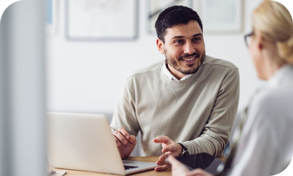 Businessperson in tan sweater sits in front of a laptop and discusses something with a colleague. 
