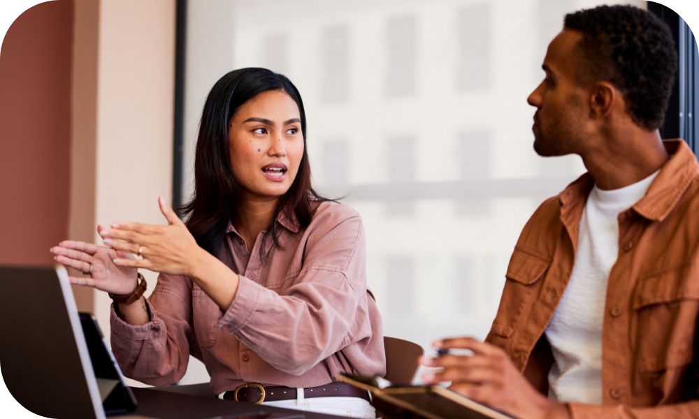 Two businesspeople in business casual dress discuss something in front of laptops. 