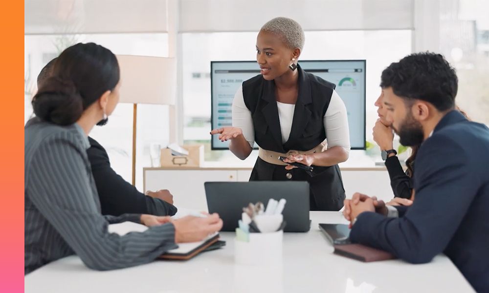 Five businesspeople are engaged in a discussion with one standing at the end of the table.