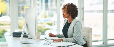 A professional sits at a desk while writing in a notebook and typing on a keyboard.