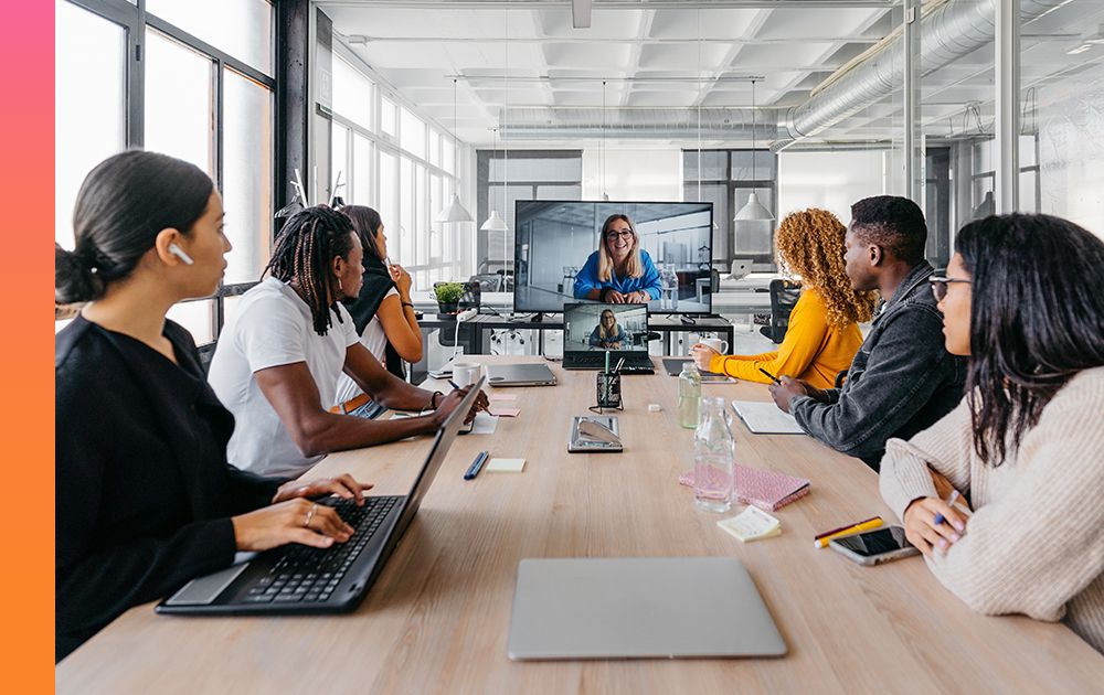 Employees sit around a conference table with a video call cast on a screen at the end of the table.