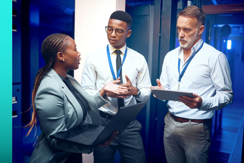 Three businesspeople holding devices and standing together and talking in front of a server room