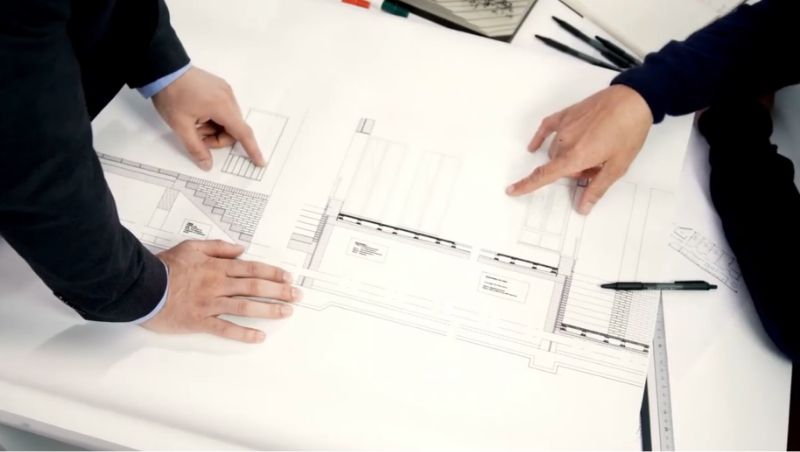  Zwei Personen sehen sich Papiere an, die an einem Schreibtisch ausgelegt sind, und zeigen auf verschiedene Abschnitte 