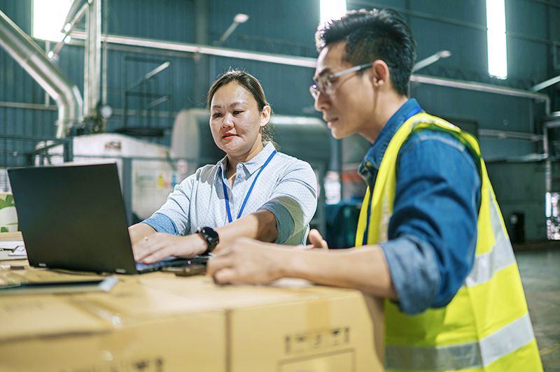 Two workers using a laptop sitting on top of a box in a warehouse