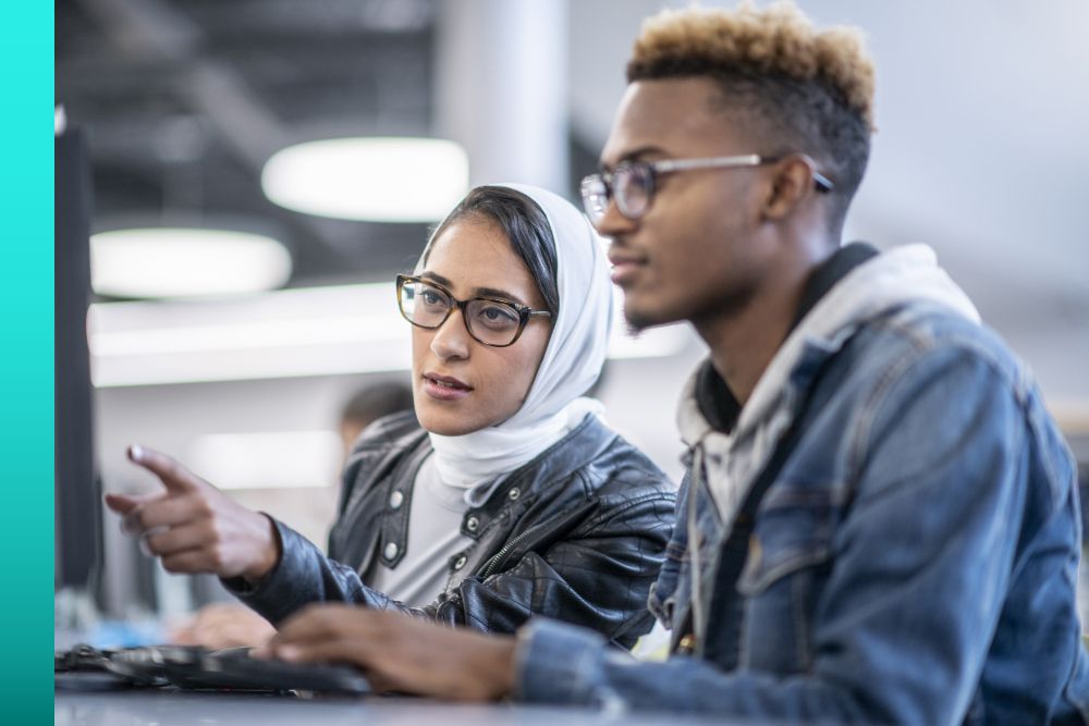 Two coworkers collaborate at a computer in a modern office, with one pointing at the screen while discussing a task.