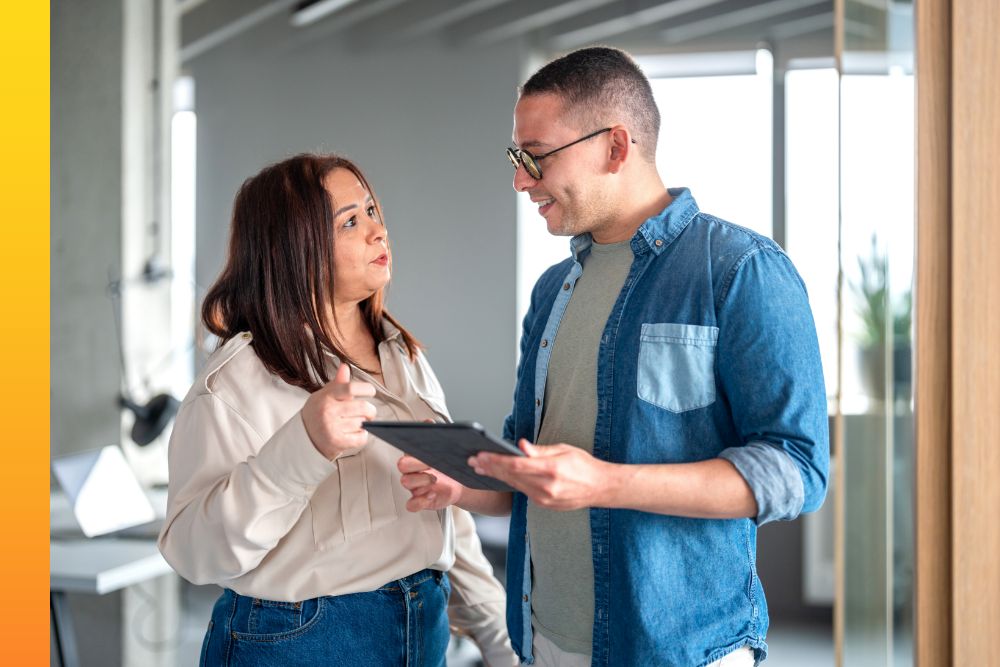 Two business people talking in an office, one holding a tablet and wearing a denim shirt.