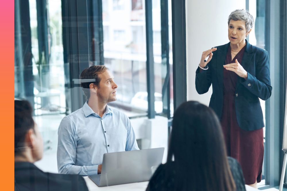 Three people sitting at a conference table while they watch another person while they're standing up and speaking