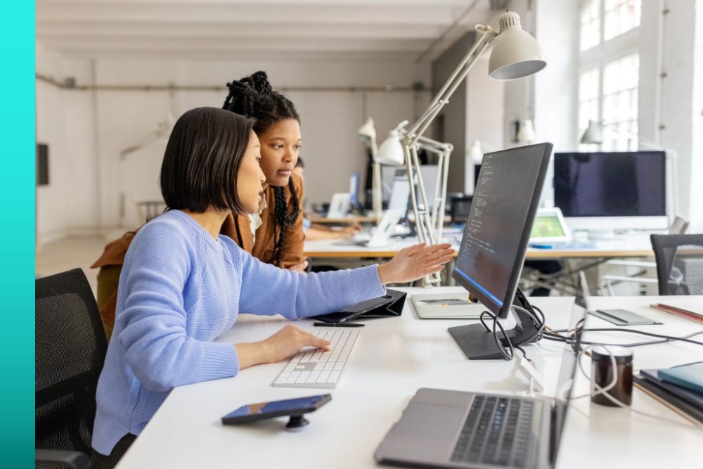 Two professionals seated at a desk in an office pointing at information displayed on a computer monitor.