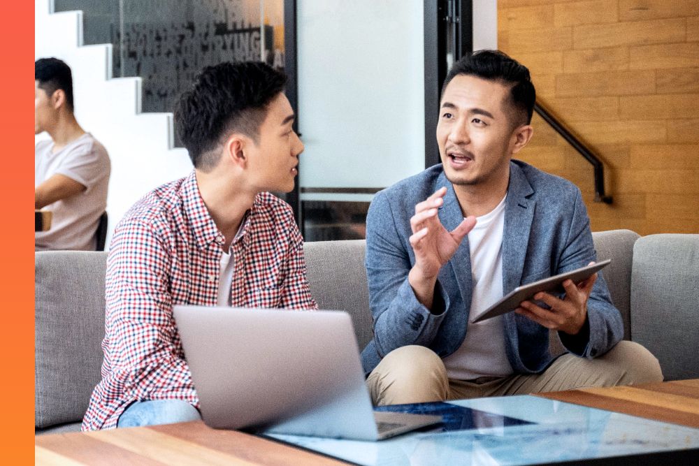 Two people sitting on a couch talking and the person on the right is holding a tablet and gesturing