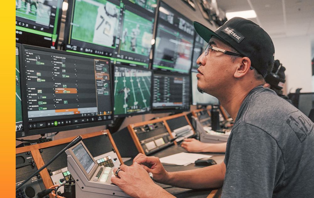 Pac-12 technician wearing a baseball cap and glasses, operating a mixing board in front of multiple monitors displaying sports footage and data.