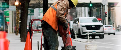 A Lumen worker in a yellow hardhat and orange safety vest drills concrete on a Chicago sidewalk with traffic and buildings in the background