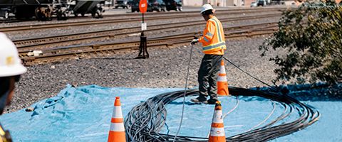 Two Lumen workers in white hardhats and orange safety vests lay fiber cable in figure-eight shape near railroad tracks