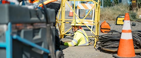 Lumen worker in yellow hardhat and safety gear lowers into a manhole near roadway to start fiber pulling