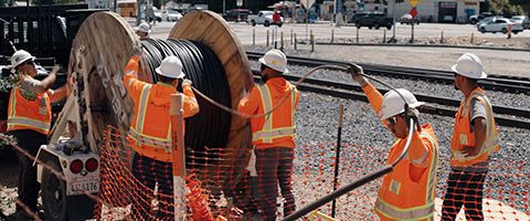 Five Lumen workers pull fiber near a railroad in white hardhats and orange safety vests