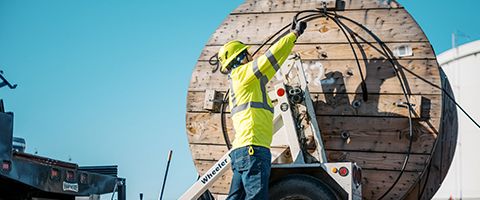 A Lumen worker in yellow hardhat and safety vest unhooks a wooden fiber roll from a utility truck