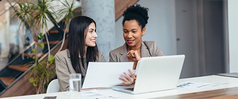 Two people smile and discuss business in front of a table with a laptop.