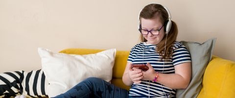 Person sitting on a couch and wearing headphones while looking down at a smart phone. 