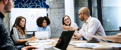 Five security specialists engaged in spirited discussion in conference room with laptops and reports strewn across the table.