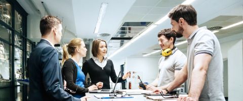 Businesspeople standing at a conference table listening to a presenter.